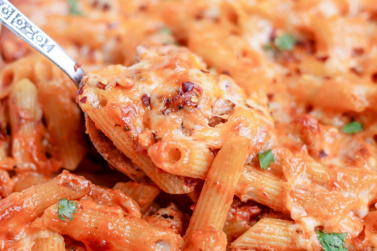 Close-up of baked penne pasta covered in melted cheese and tomato sauce, with a serving spoon lifting a portion.