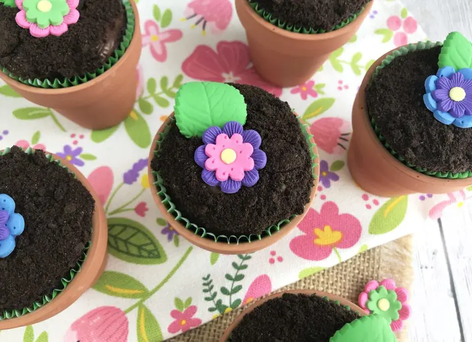 Cupcakes in small flowerpots topped with crushed cookie โsoilโ and decorated with colorful, edible flower and leaf shapes, placed on a floral-patterned cloth.