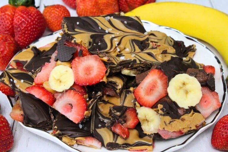 A plate of chocolate bark swirled with peanut butter, topped with sliced strawberries and bananas, surrounded by fresh strawberries and a banana.