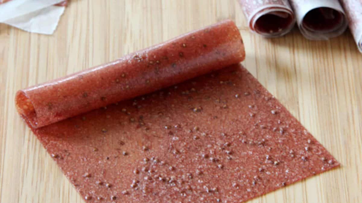 A sheet of homemade fruit leather partially rolled up on a wooden surface, with additional rolled fruit leathers in the background.