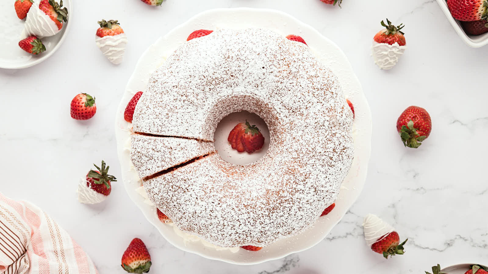 A round bundt cake dusted with powdered sugar sits on a white plate, garnished with fresh strawberries and surrounded by more strawberries on a marble surface.