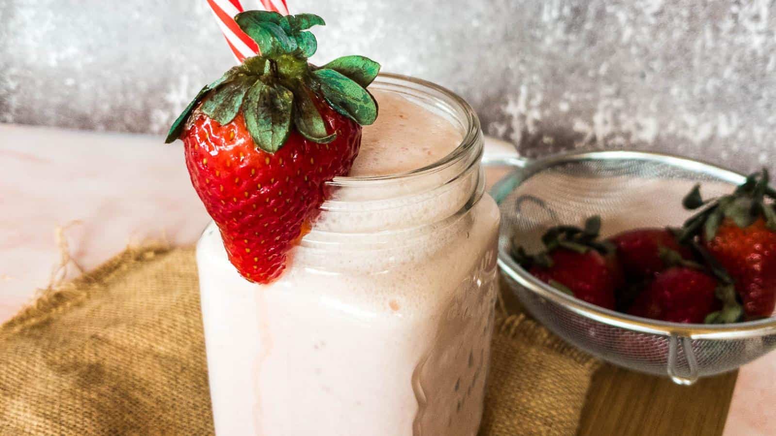 A mason jar filled with strawberry smoothie, topped with a large fresh strawberry, sits on burlap beside a strainer of whole strawberries.
