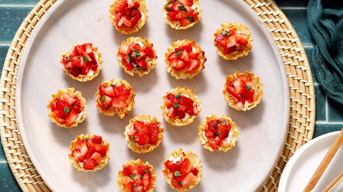 A plate of small phyllo pastry cups filled with diced strawberries and garnished with chopped herbs, arranged on a woven placemat.