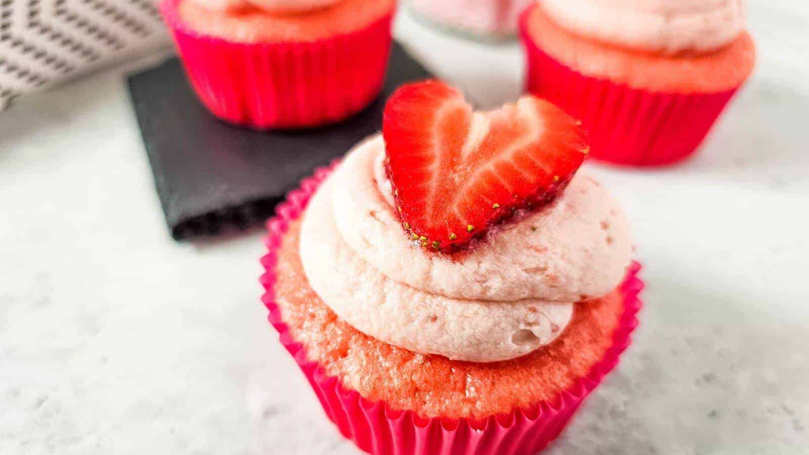 A strawberry cupcake with pink frosting is topped with a halved strawberry, and is placed in a pink wrapper. Two more cupcakes are in the background.