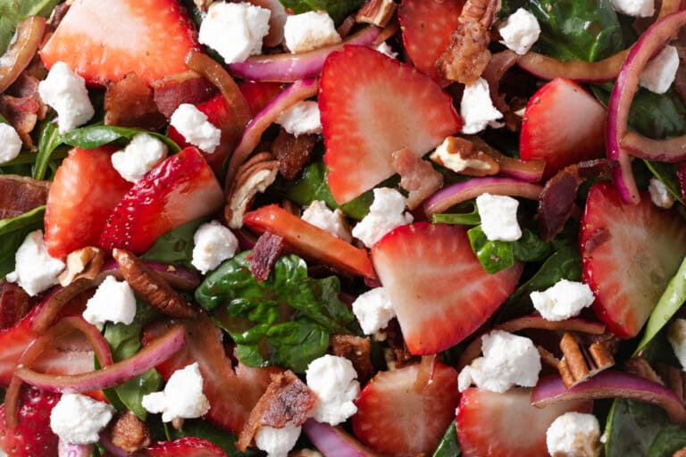 Close-up of a salad with fresh spinach, sliced strawberries, red onions, pecans, crumbled goat cheese, and pieces of bacon.