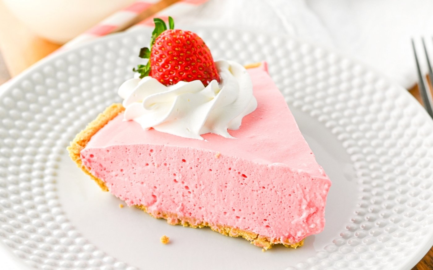 A slice of pink strawberry cream pie with a graham cracker crust, topped with whipped cream and a fresh strawberry, served on a white plate.