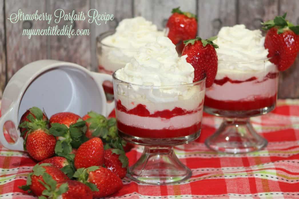 Three glass dishes of strawberry parfait topped with whipped cream and a whole strawberry, with fresh strawberries and a measuring cup beside them on a red plaid cloth.