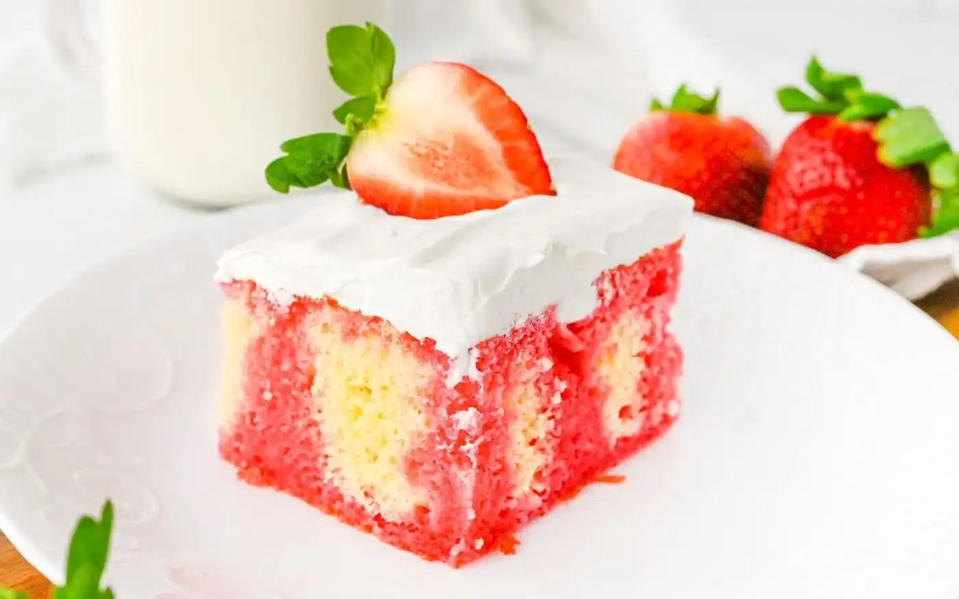 A slice of strawberry poke cake with white frosting and a halved strawberry on top, served on a white plate with whole strawberries in the background.