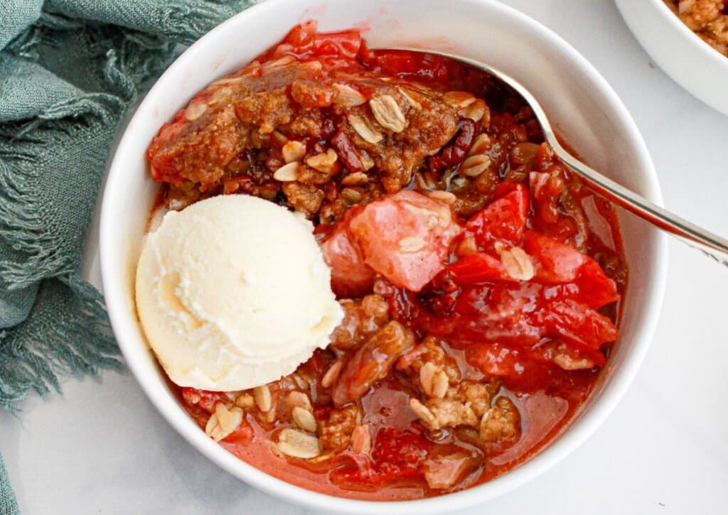 A bowl of strawberry crisp topped with a scoop of vanilla ice cream, featuring oat and brown sugar crumble, with a spoon inside the bowl.