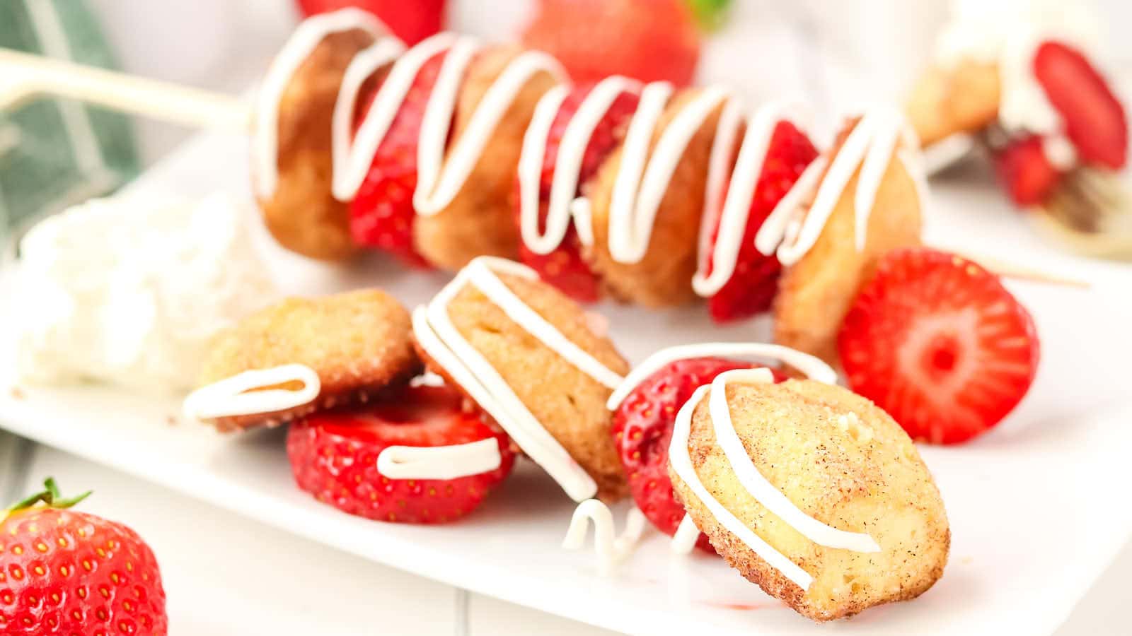 A skewer with alternating mini donuts and strawberry slices, drizzled with white icing, is displayed on a white plate with extra strawberries and donuts.