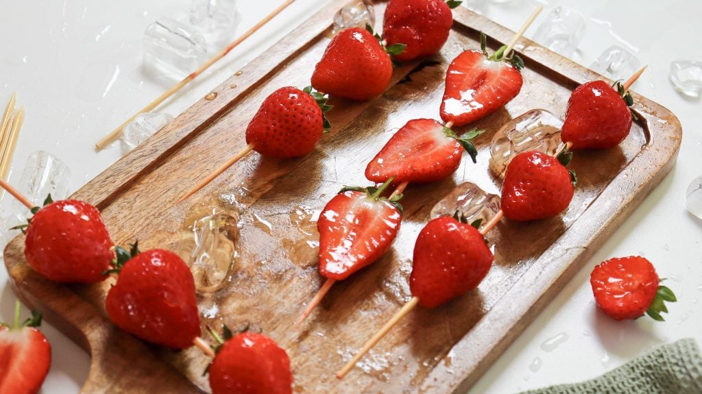 Whole and halved strawberries and ice cubes are skewered on wooden sticks and placed on a wooden cutting board.