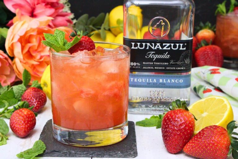 A glass of strawberry cocktail with ice and mint garnish sits beside a Lunazul Tequila Blanco bottle, surrounded by strawberries, lemon, and flowers.