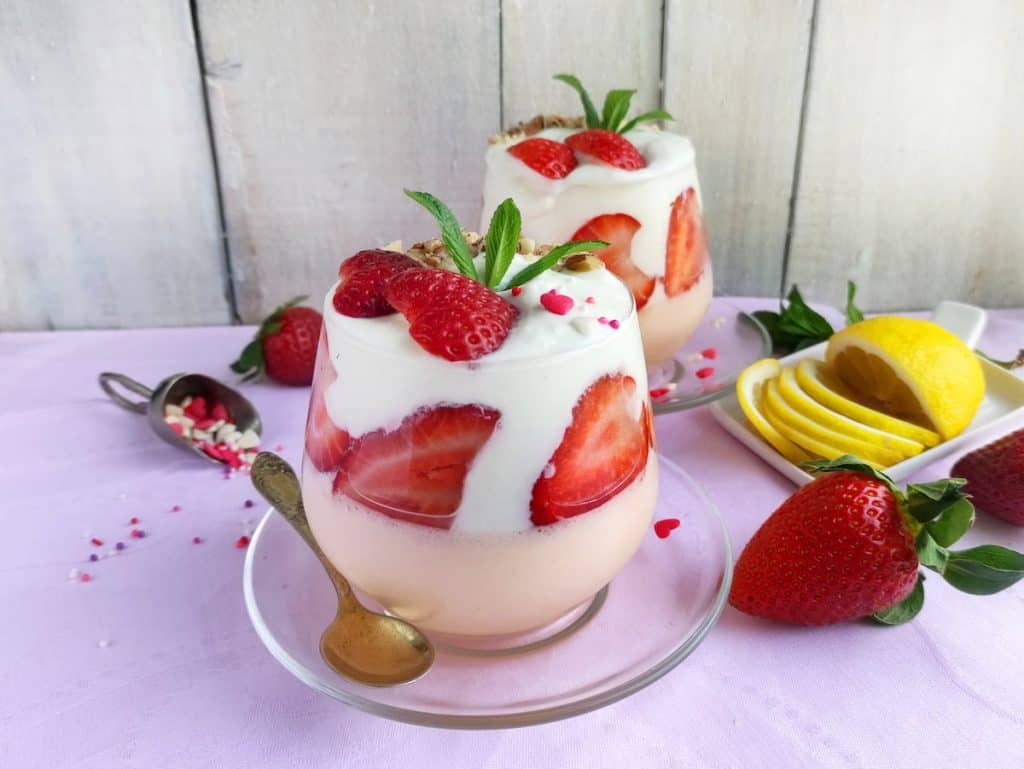Two glasses of layered strawberry parfait with cream, fresh strawberries, and mint, served on saucers with spoons, next to sliced lemon and whole strawberries on a pink cloth.
