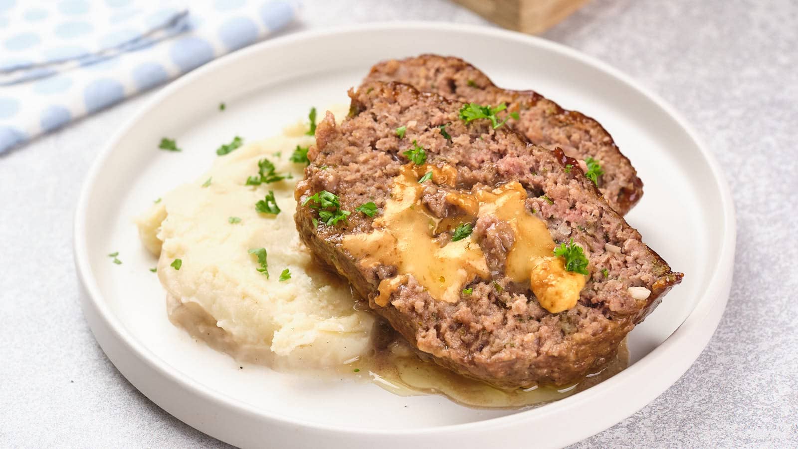 A plate with two slices of meatloaf topped with gravy, served alongside a portion of mashed potatoes and garnished with chopped parsley.