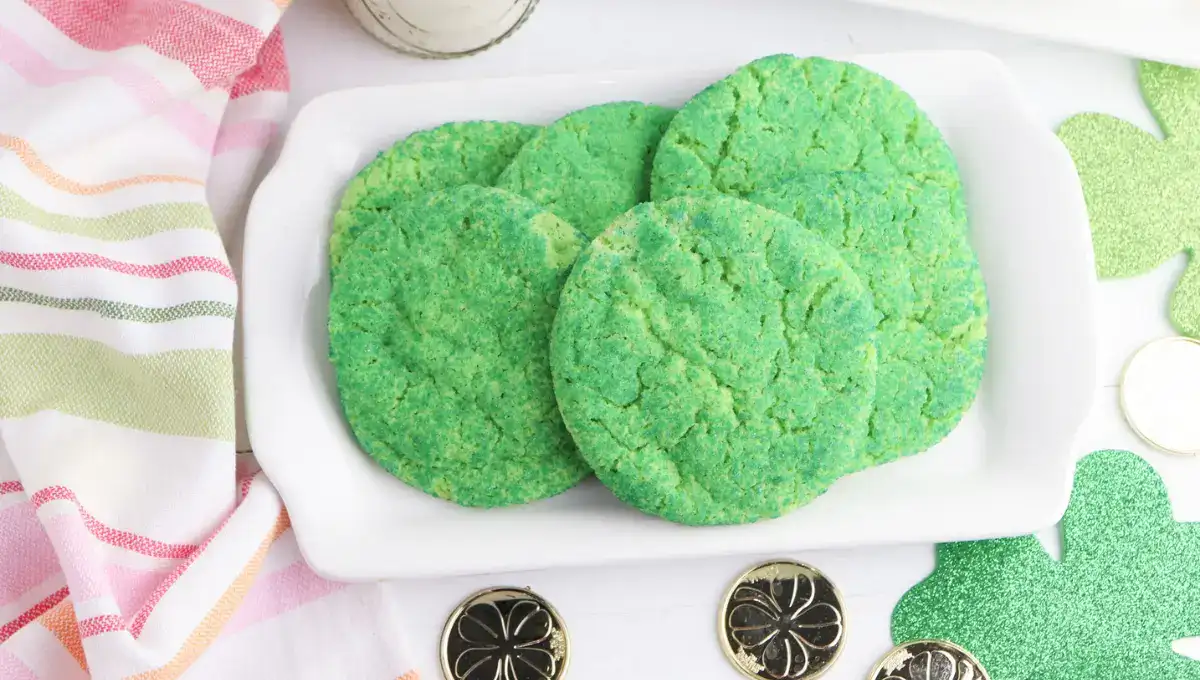 Four green cookies are arranged on a rectangular white plate, placed on a white surface with a striped cloth and gold coins nearby.