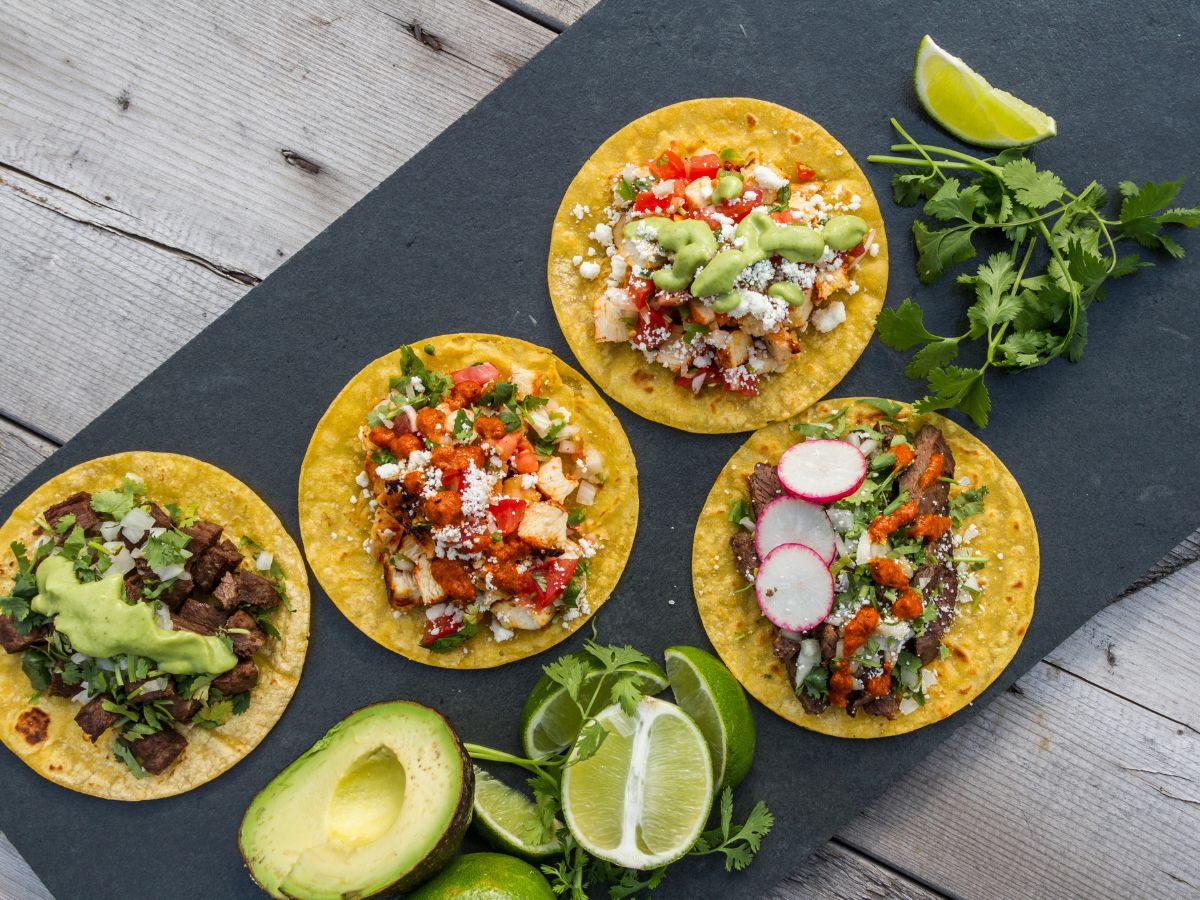 Four assorted tacos topped with various vegetables, cheese, and sauces are arranged on a slate board with lime wedges, cilantro, and halved avocado nearby.