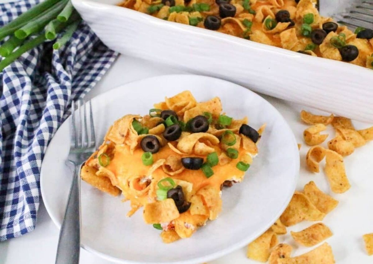 A serving of Frito pie topped with melted cheese, black olives, and green onions on a white plate, with a fork and a casserole dish of Frito pie in the background.