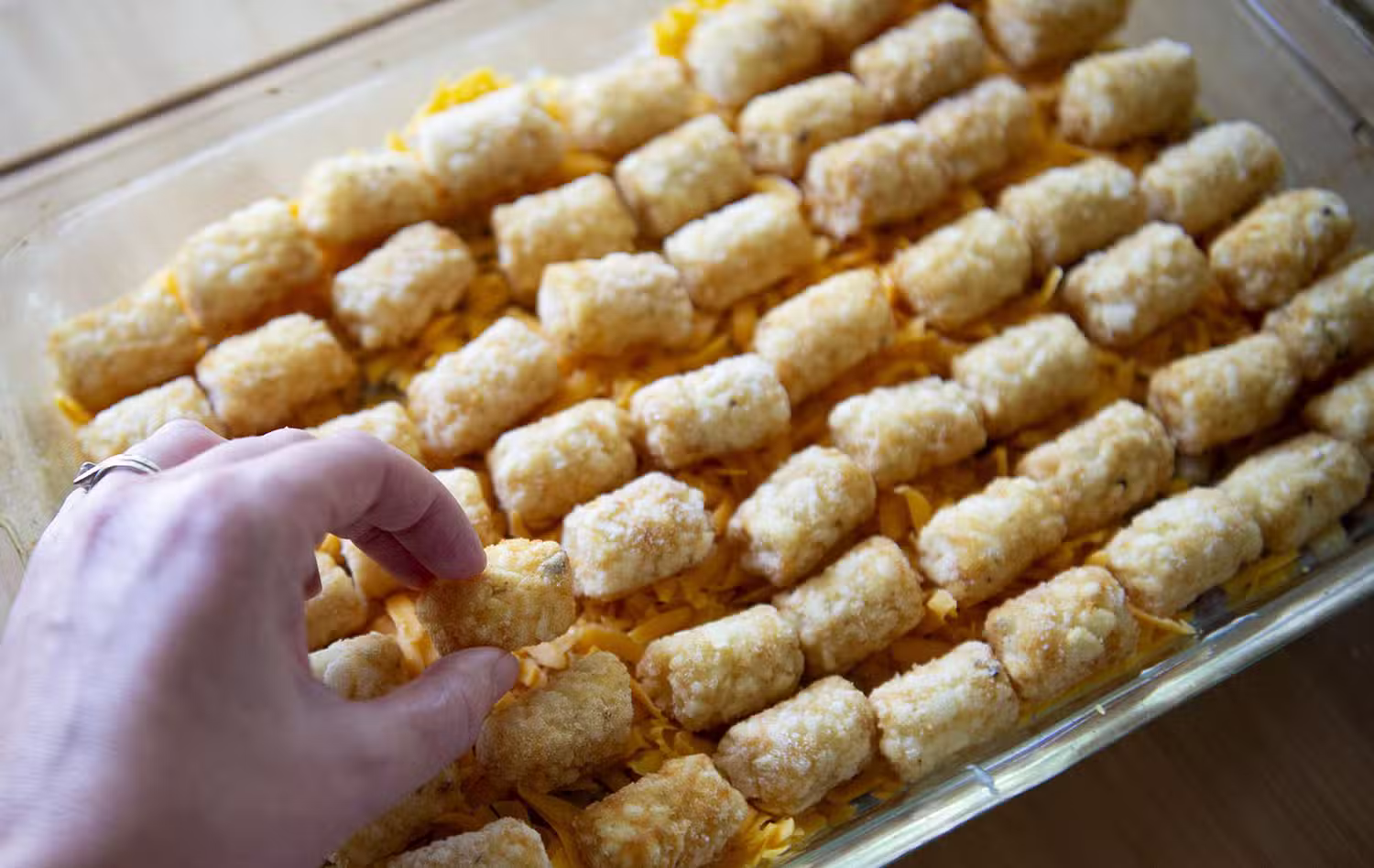A hand places tater tots in neat rows over shredded cheese in a glass baking dish on a wooden surface.