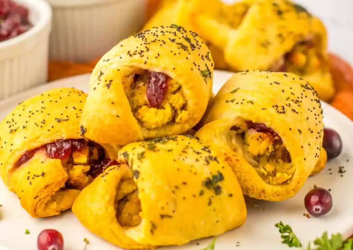 Four golden crescent rolls filled with turkey, stuffing, and cranberry sauce are arranged on a white plate, garnished with herbs and poppy seeds.