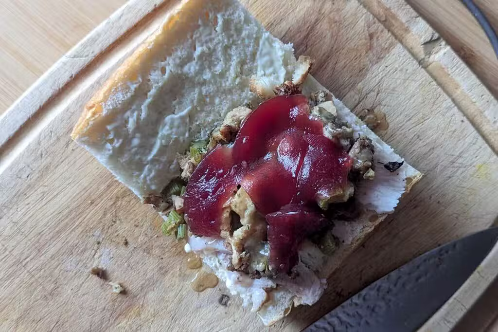 A slice of bread with stuffing, cranberry sauce, and a white spread on a wooden cutting board next to a knife.