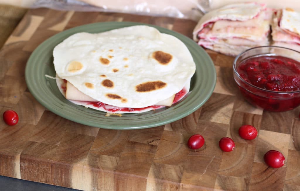 A tortilla wrap with turkey, cheese, and cranberry sauce on a green plate, with a bowl of cranberry sauce and whole cranberries on a wooden cutting board.