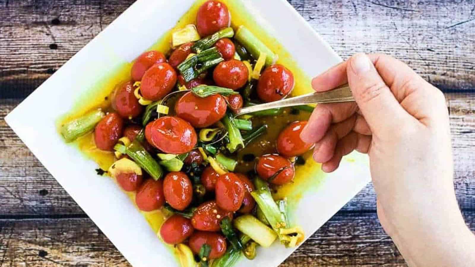 A hand holds a fork over a square white plate filled with cherry tomatoes, scallions, and yellow sauce on a wooden surface.