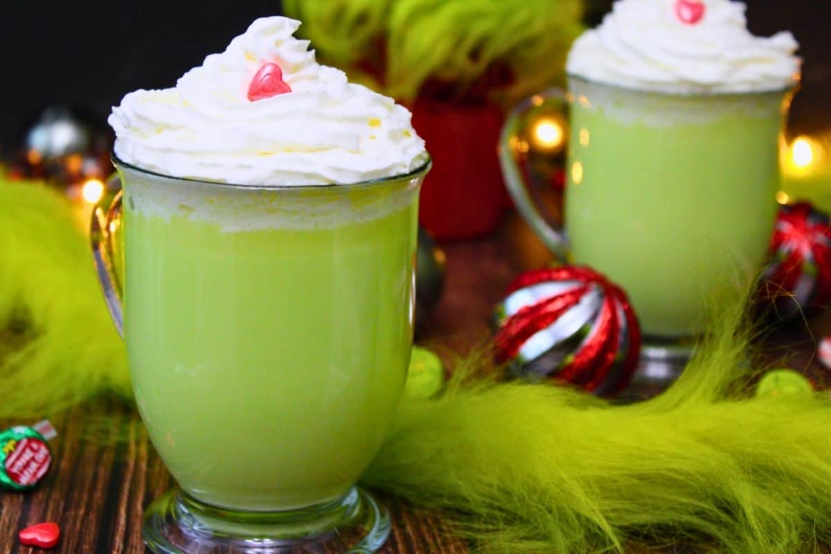 Two glass mugs filled with light green beverage topped with whipped cream and a small red candy, surrounded by green fur-like material and holiday decorations.