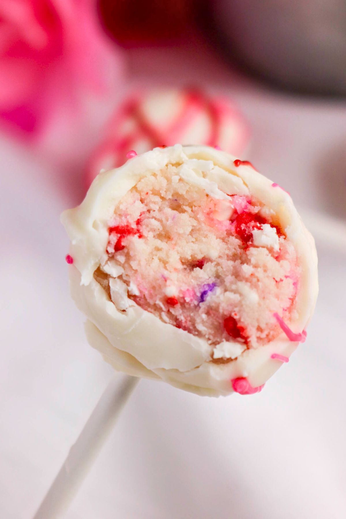 A close-up of a white chocolate-covered cake pop with a pink and red sprinkle-filled cake center on a white stick.