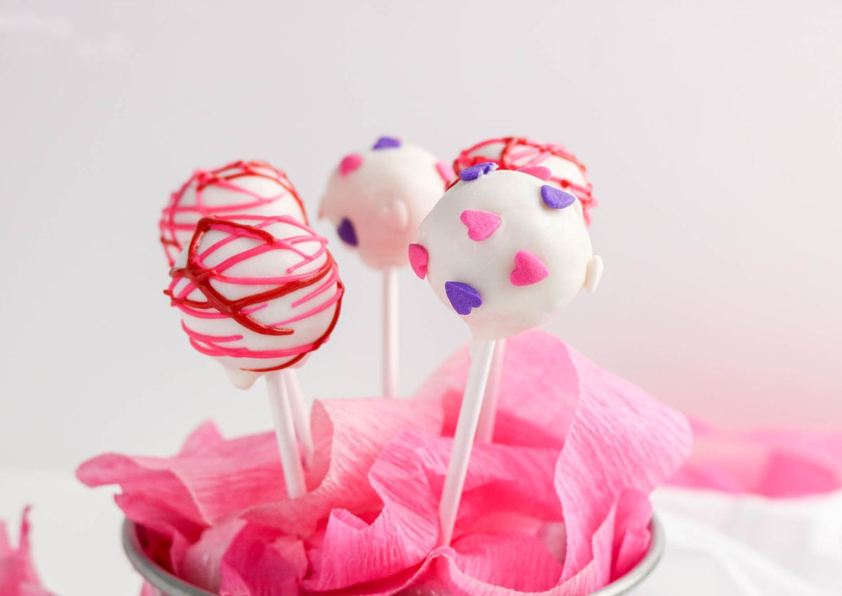 Four white cake pops on sticks, decorated with pink, red, and purple heart sprinkles and red icing, placed in pink tissue paper inside a container.