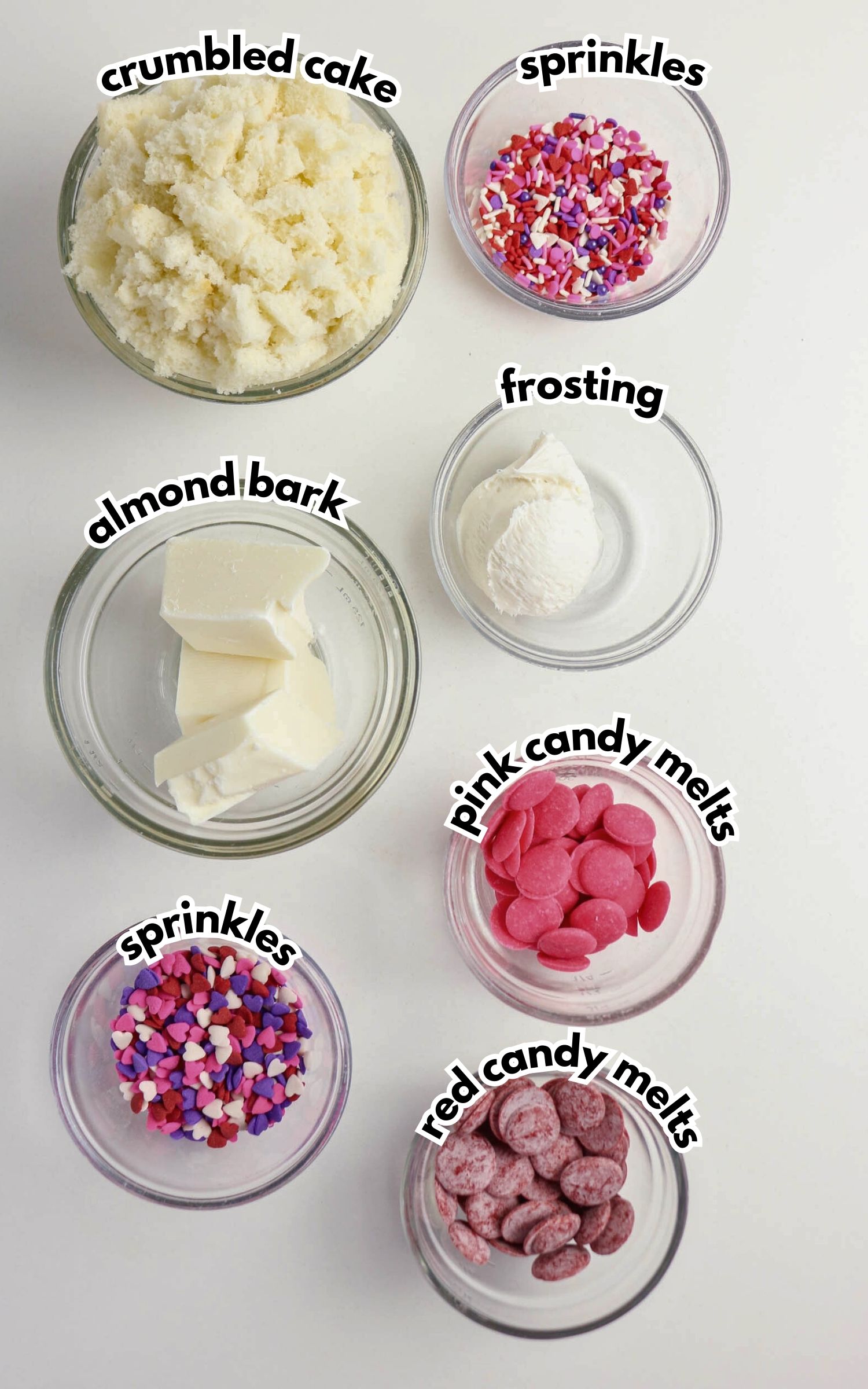 Six glass bowls containing crumbled cake, almond bark, frosting, pink candy melts, red candy melts, and two types of sprinkles, labeled accordingly.