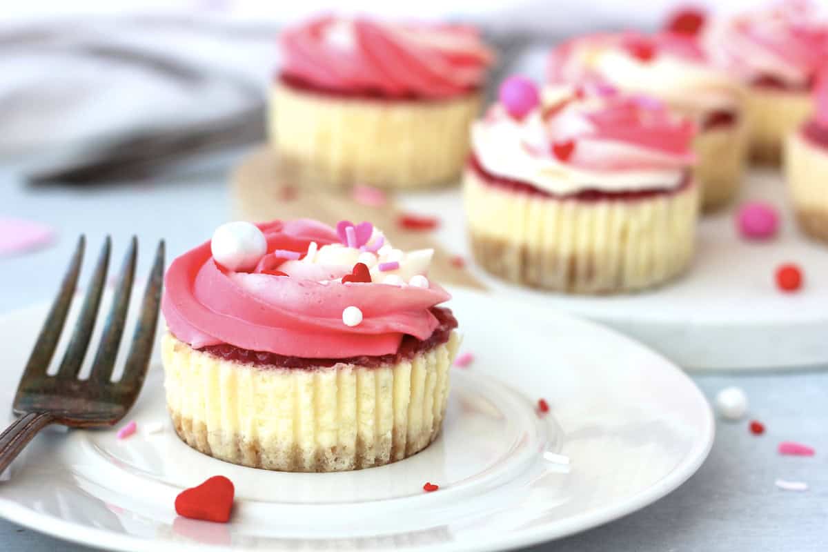 A cheesecake cupcake with pink frosting and heart-shaped sprinkles sits on a white plate next to a fork, with more cupcakes in the background.