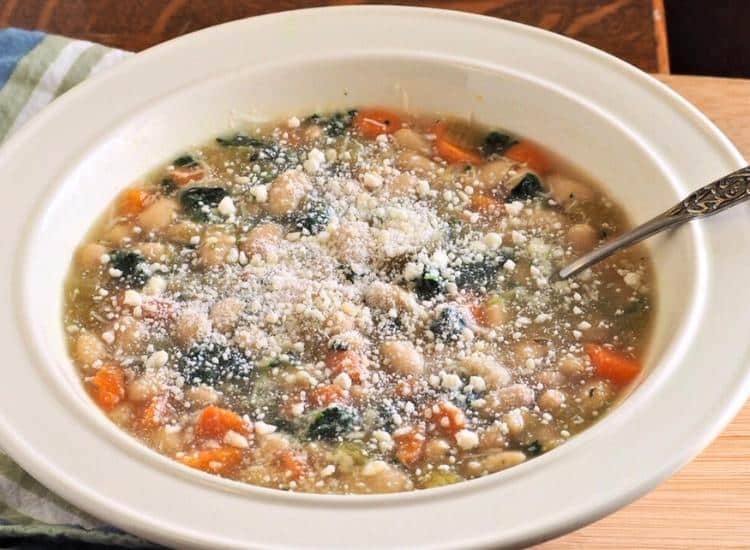 A bowl of white bean soup with carrots, greens, and grated cheese on top, with a spoon in the bowl.
