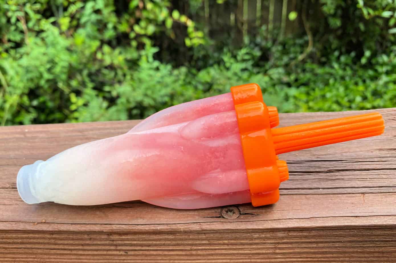 A red and white homemade popsicle with an orange handle rests on a wooden surface, with greenery visible in the background.
