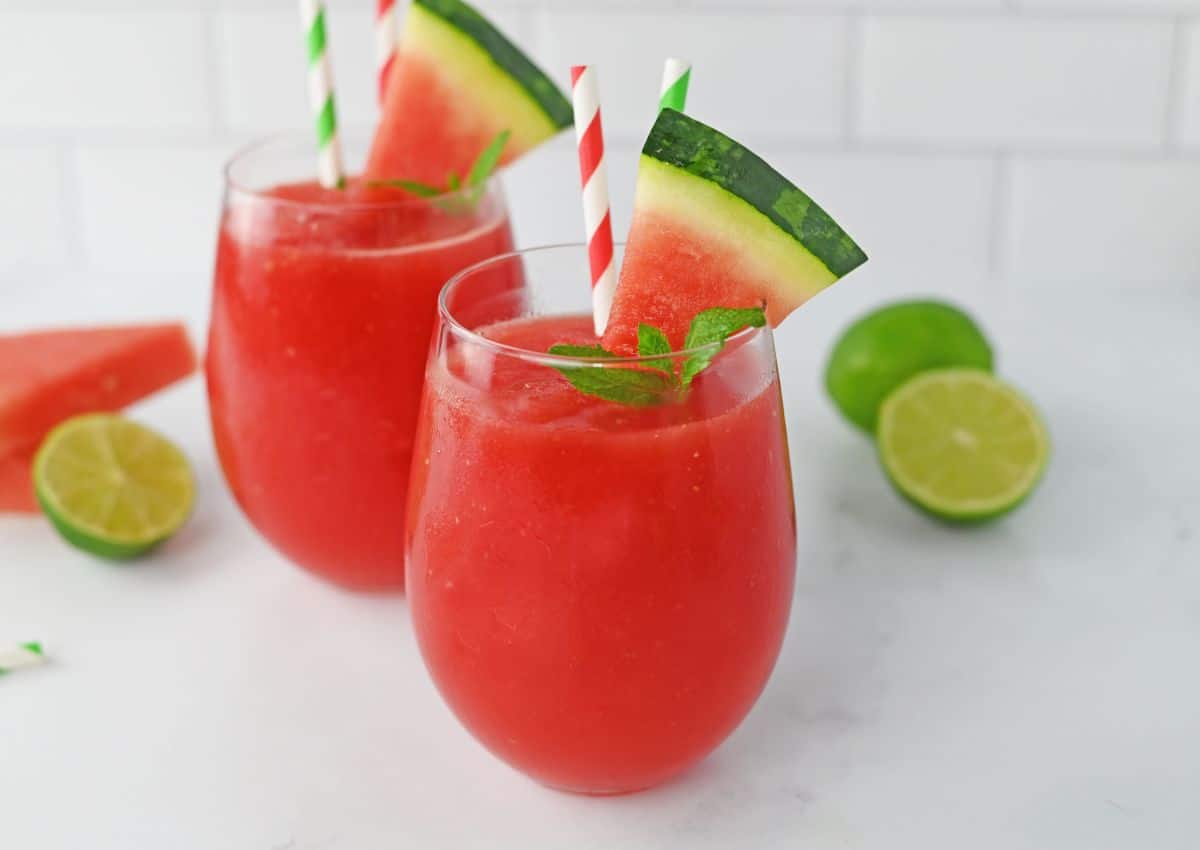 Two glasses of red watermelon drink with ice, garnished with watermelon slices, mint leaves, and striped straws. Whole and halved limes are in the background on a white surface.
