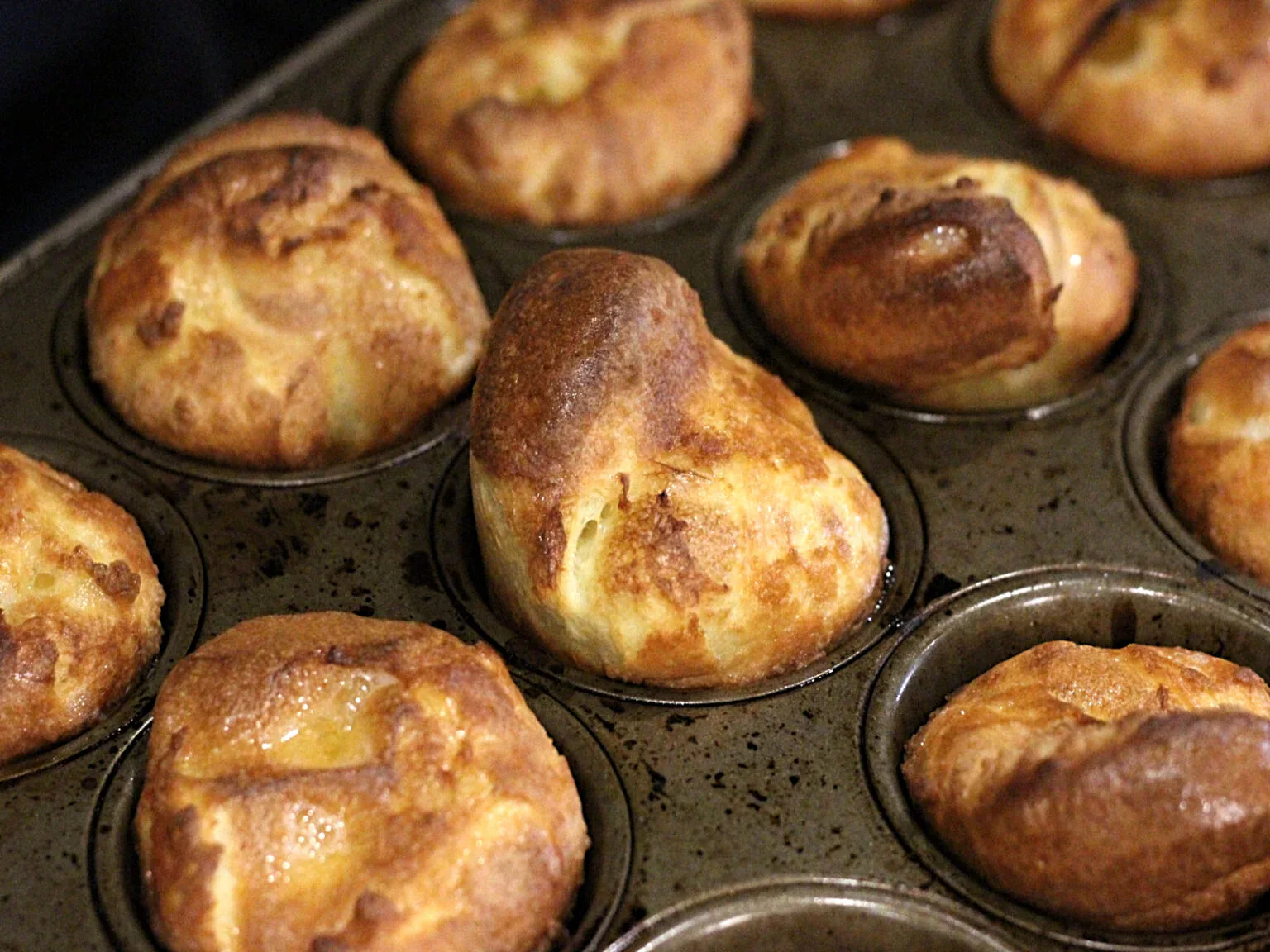 A close-up of freshly baked Yorkshire puddings in a metal muffin tin, showing golden brown, puffed-up tops.