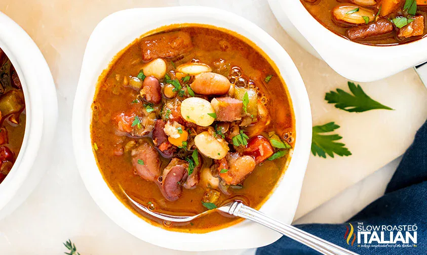 A bowl of mixed bean soup with vegetables and herbs, served on a white surface with a spoon and garnished with parsley.