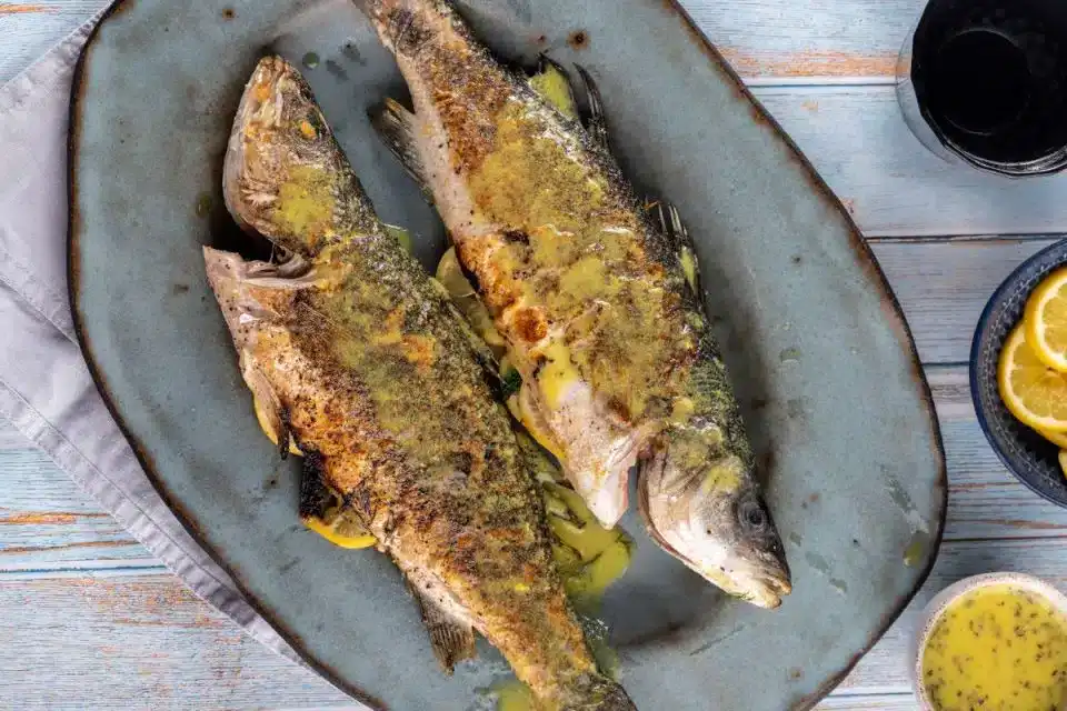 Two whole cooked fish with herbs and yellow sauce served on a gray oval plate, with lemon slices and sauce on the side.