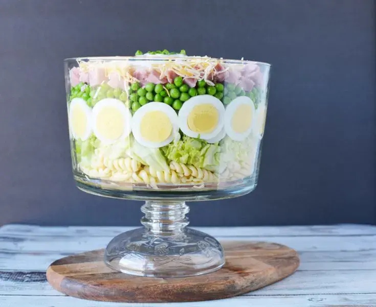 A layered salad in a glass trifle bowl with sliced eggs, pasta, lettuce, peas, ham, and shredded cheese, placed on a wooden board against a dark background.