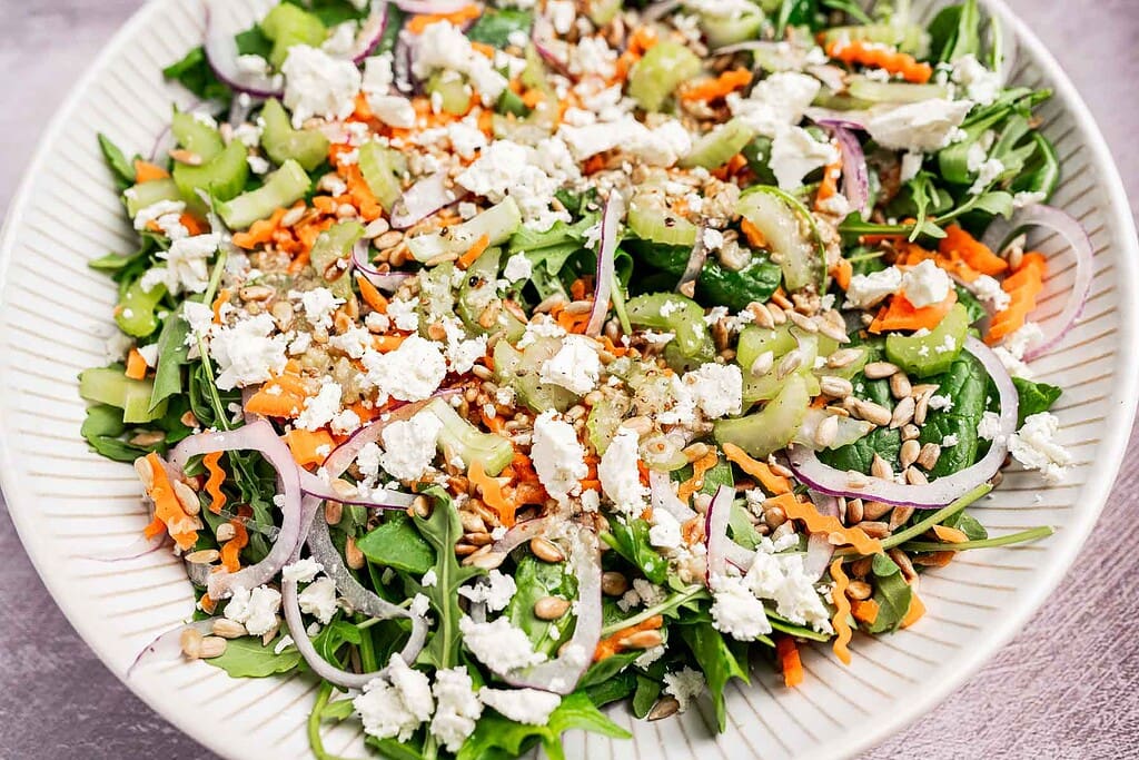 A plate of salad with arugula, shredded carrots, sliced celery, red onion, sunflower seeds, crumbled feta cheese, and a light dressing.