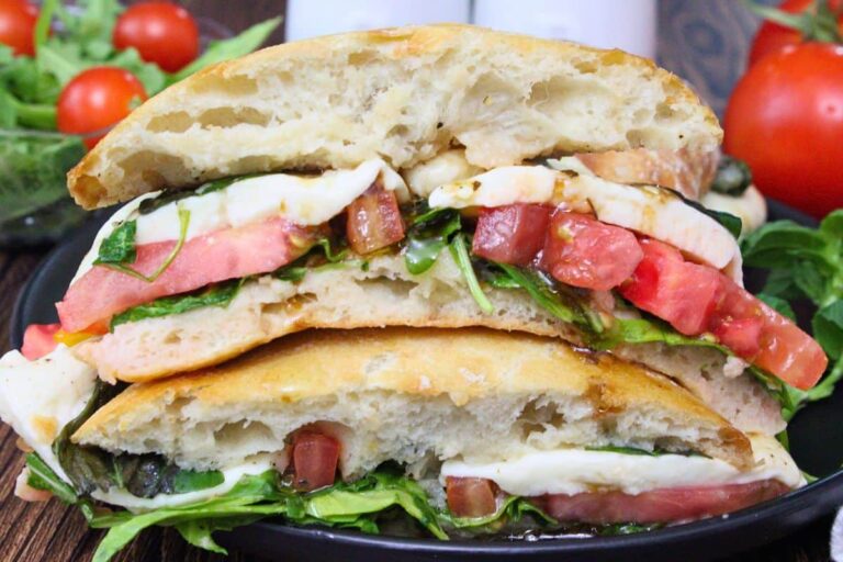 A ciabatta sandwich filled with fresh mozzarella, tomato slices, green leaves, and drizzled with balsamic glaze, stacked on a black plate with tomatoes and greens in the background.