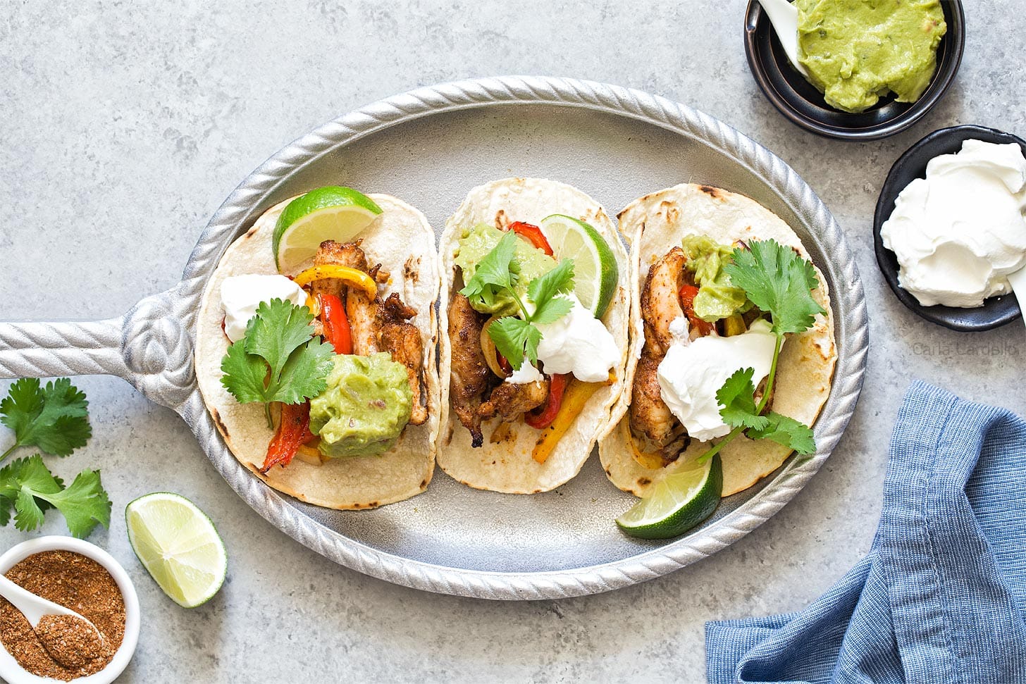 Three tacos with meat, grilled vegetables, guacamole, sour cream, and cilantro on a platter, with lime wedges, seasoning, and small bowls of guacamole and sour cream nearby.