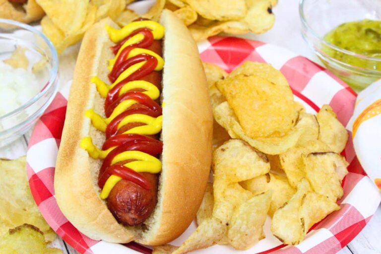A hot dog with ketchup and mustard on a bun, served on a red-and-white plate with potato chips. Guacamole and onions are in bowls nearby.