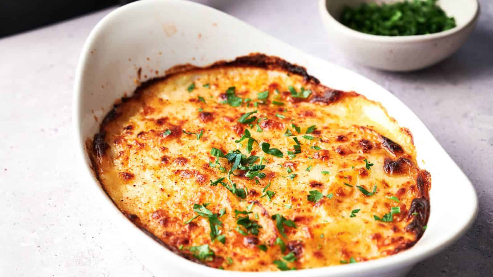 A baked dish of cheesy potato gratin topped with chopped parsley, served in a white oval casserole dish on a light surface. A bowl of chopped herbs sits in the background.
