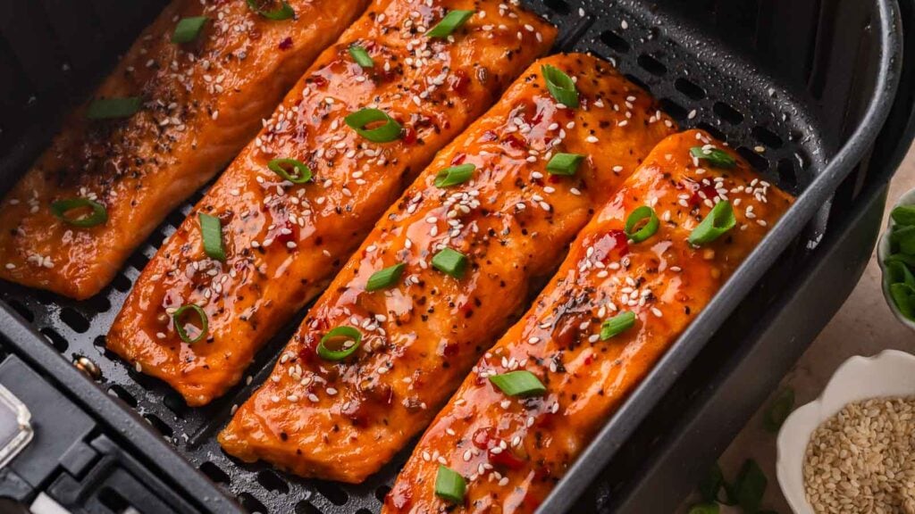 Four glazed salmon fillets topped with sesame seeds and chopped green onions cooking in an air fryer basket.