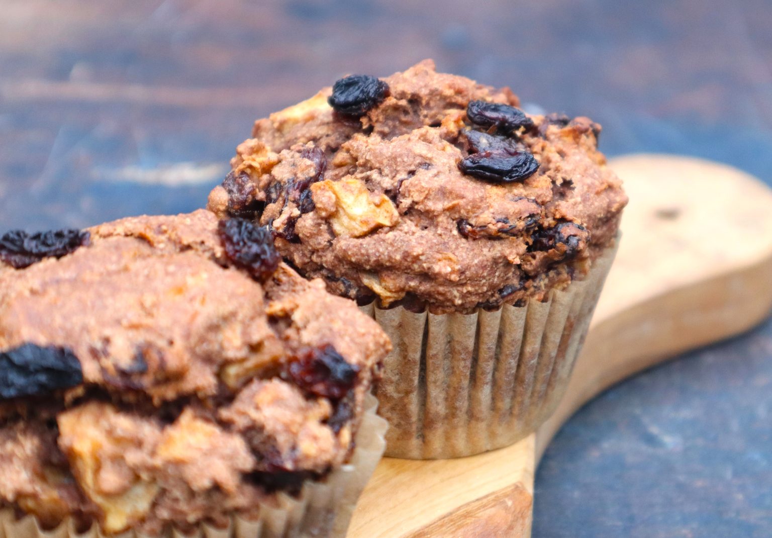 Two bran muffins with raisins and apple pieces sit on a wooden board against a blurred dark background.