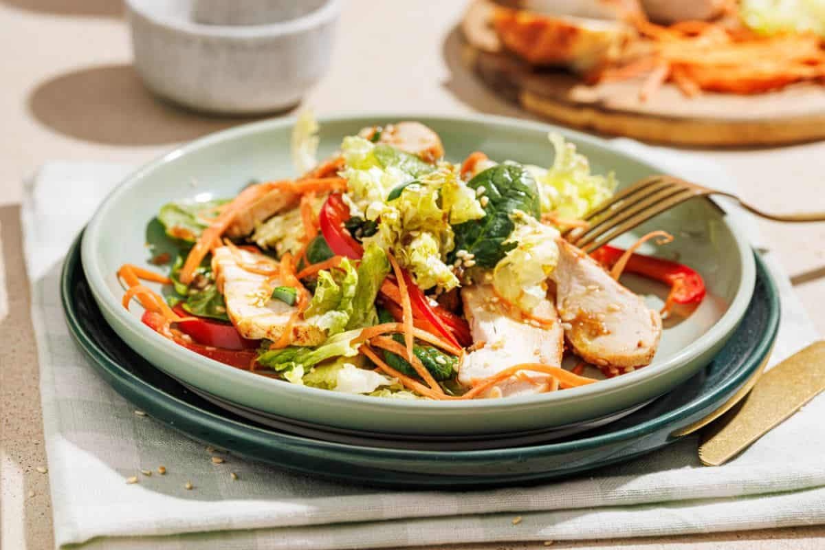 A plate of salad with sliced grilled chicken, mixed greens, shredded carrots, and red bell peppers, served with a gold fork and knife on a light table.