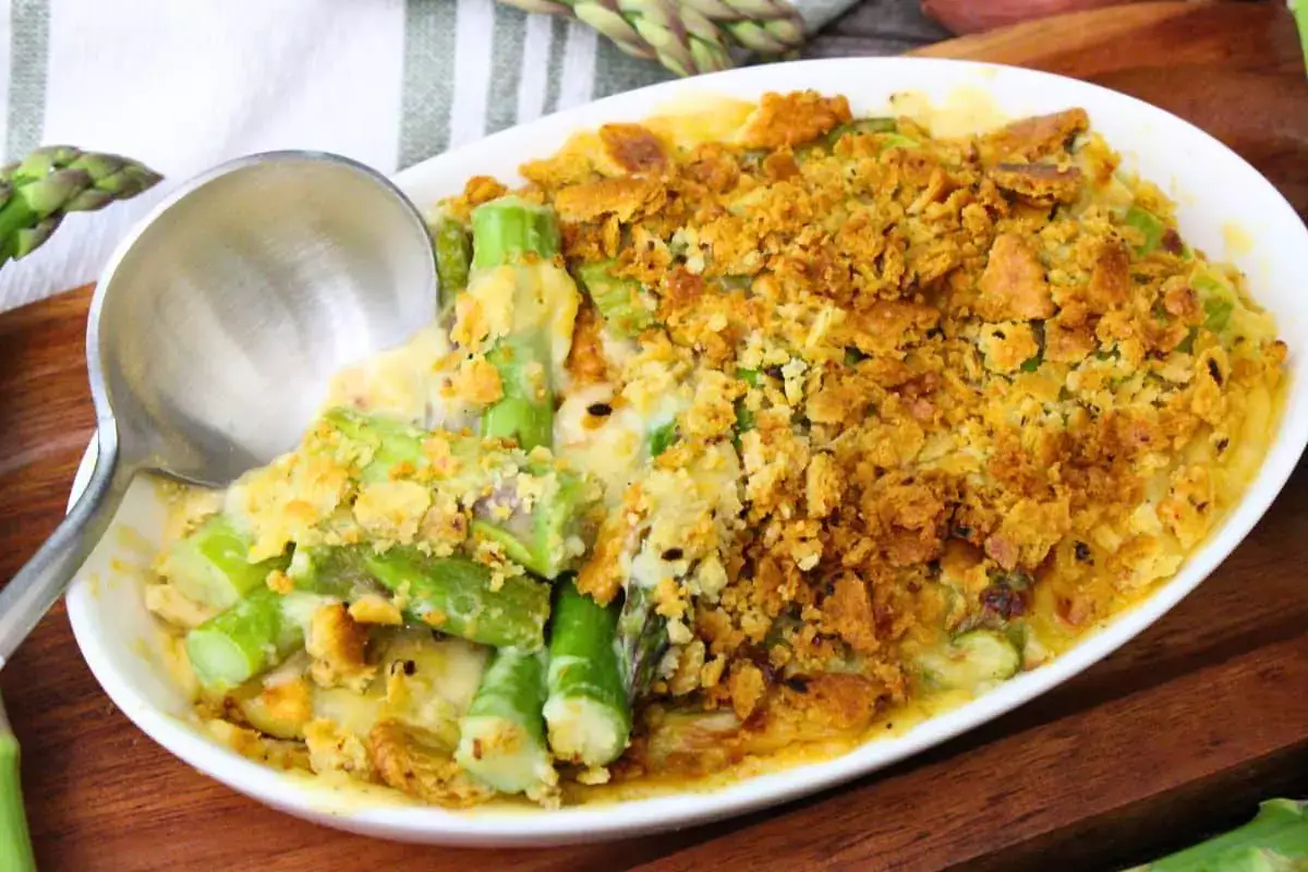 Oval dish of baked asparagus casserole topped with golden breadcrumbs, partially served with a metal spoon on a wooden surface.