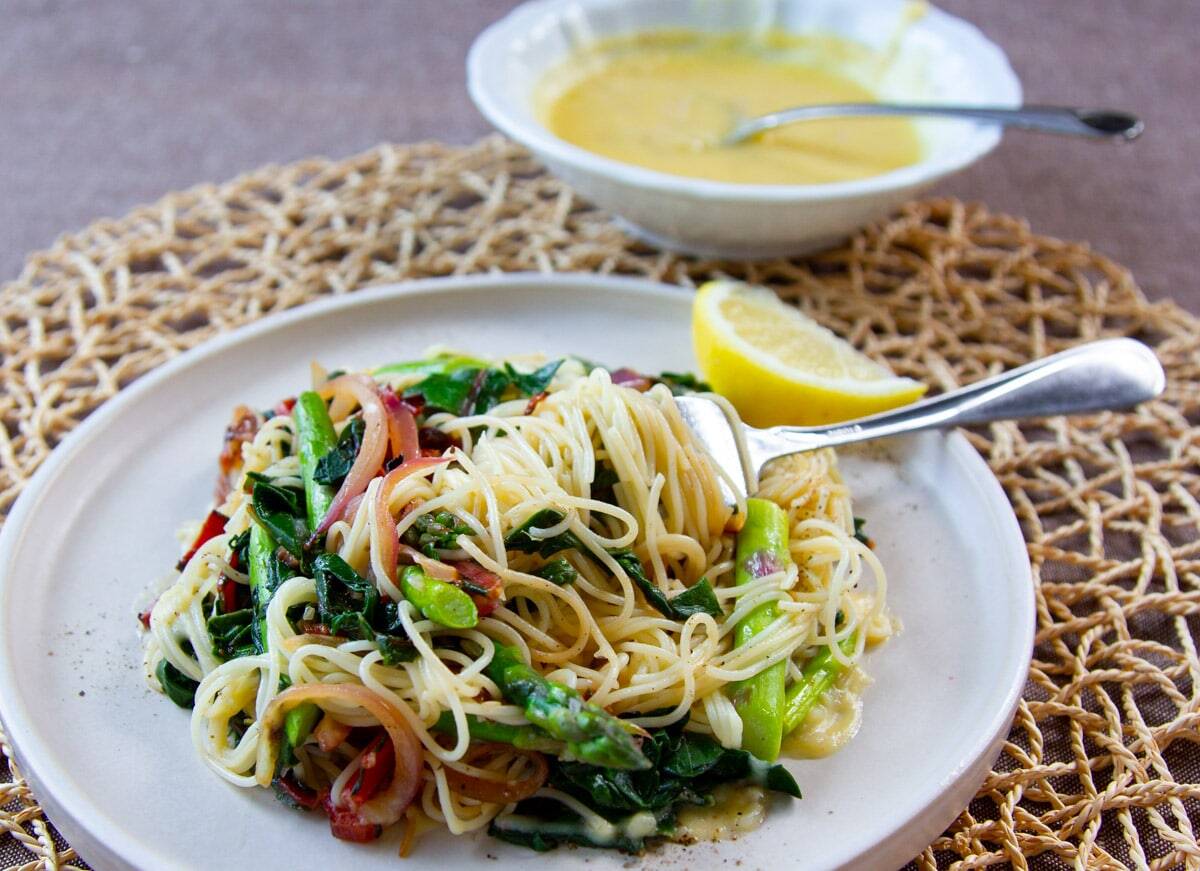 A plate of pasta with sautéed vegetables and a lemon wedge, a fork resting on top, and a bowl of yellow sauce in the background.