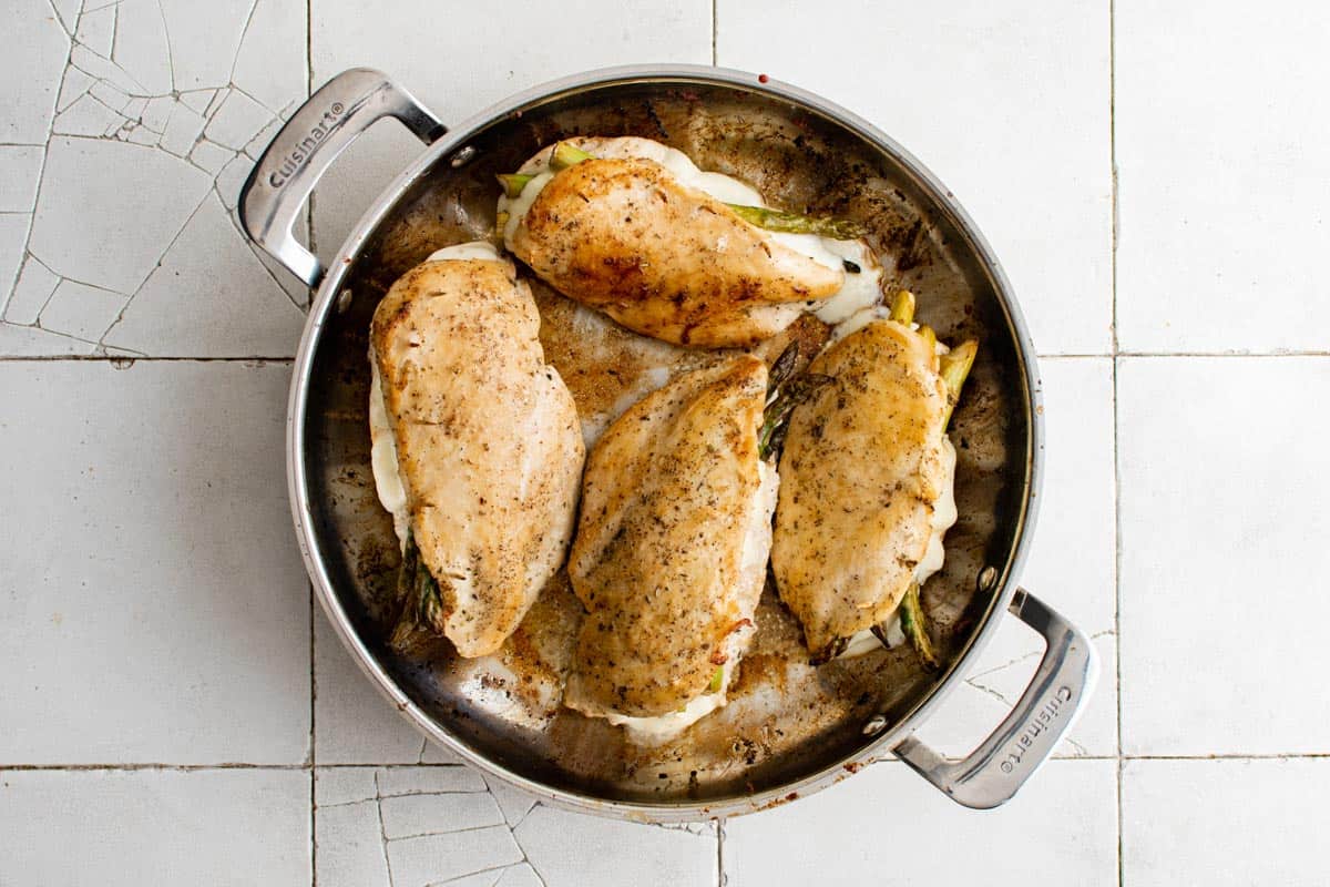 Four seasoned, stuffed chicken breasts cooking in a stainless steel Cuisinart pan on a white tiled surface.