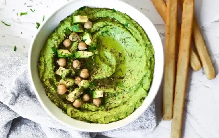 A bowl of green hummus topped with chickpeas, avocado cubes, herbs, and a drizzle of olive oil, with breadsticks on the side.